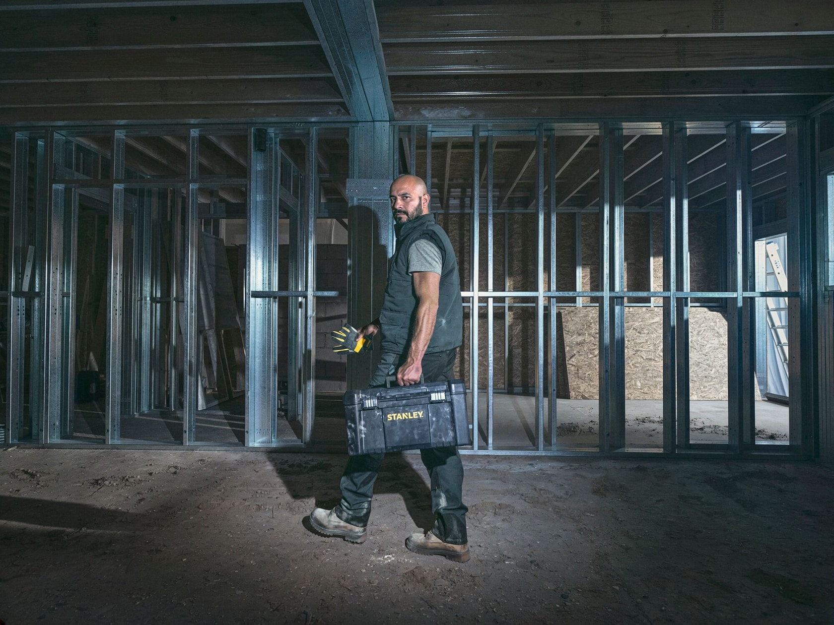 Foto de un hombre con caja de herramientas STANLEY y herramientas en una obra con estructura metálica, vista lateral.